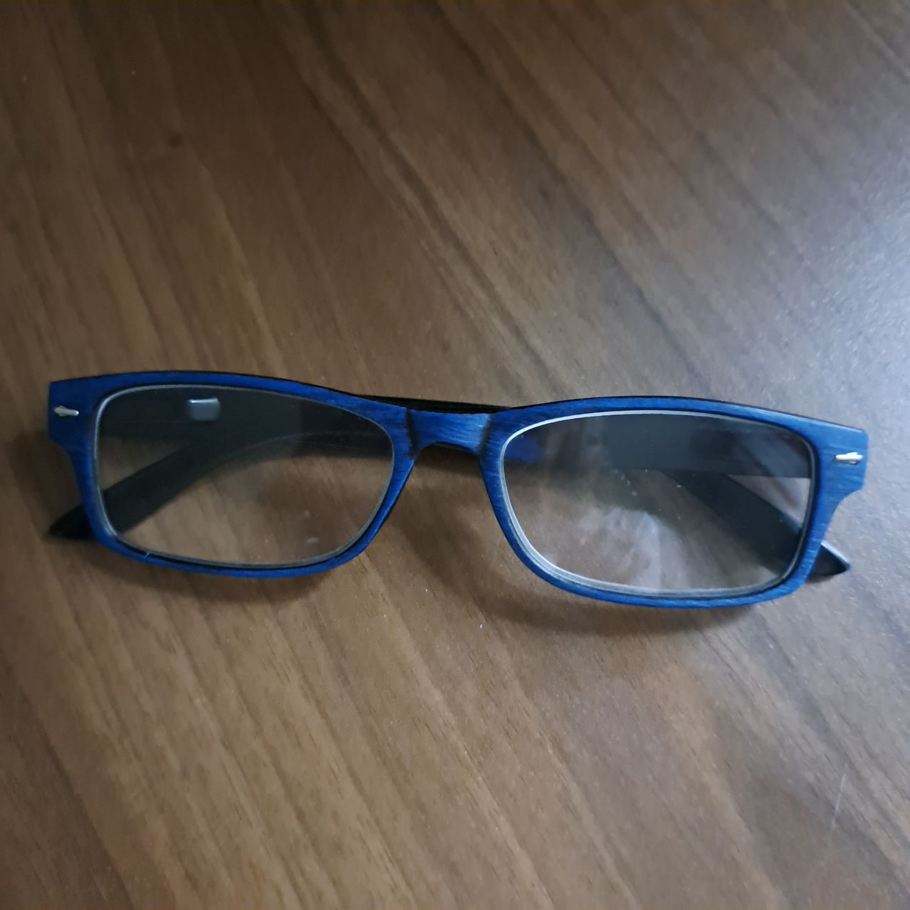 A pair of blue-framed glasses with clear lenses lies on a wooden surface. The frame appears sturdy and well-maintained.