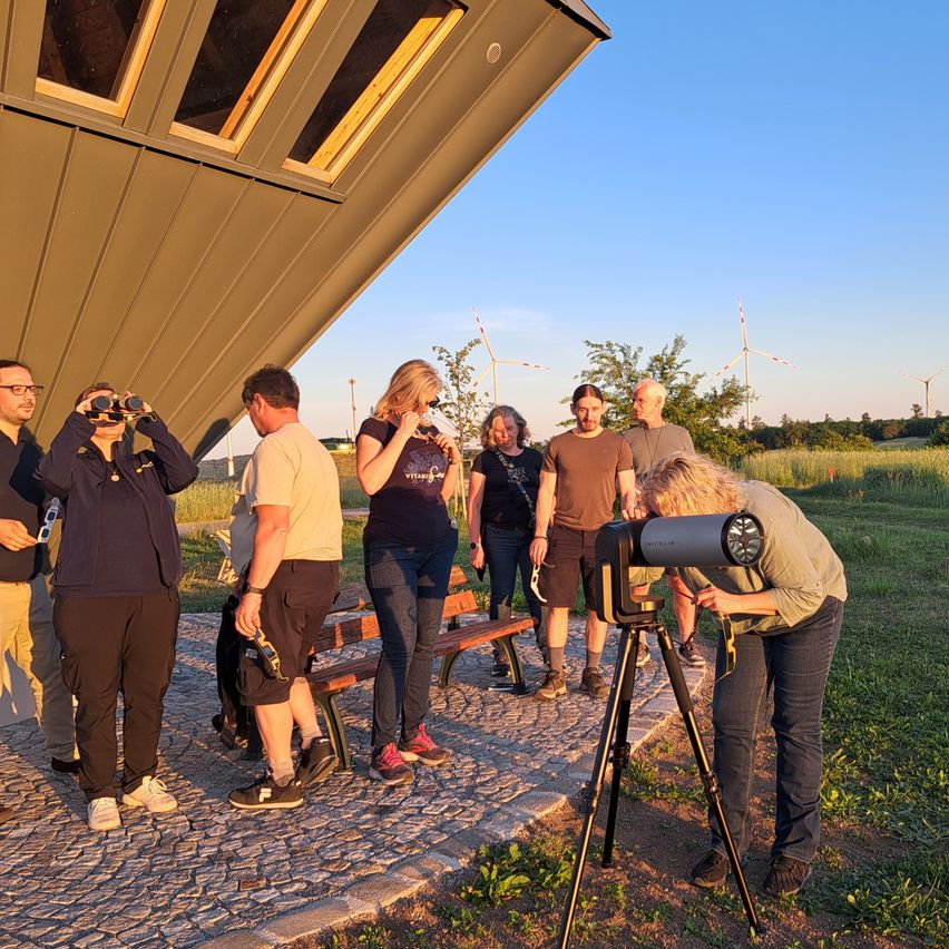 A group of people are gathered outside using telescopes to observe the sky. They are near a building with windows, a bench, and a clear view of the horizon. The sky is clear and the sun is setting.