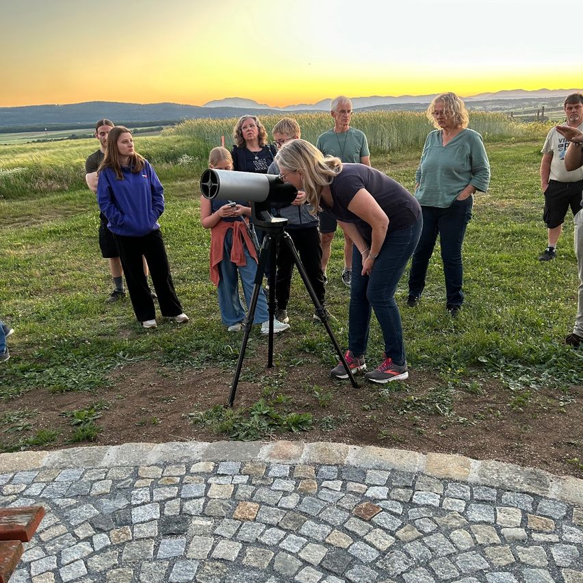 A group of people observe the sunset with a telescope on a grassy field. One woman is looking through the telescope while others stand around.