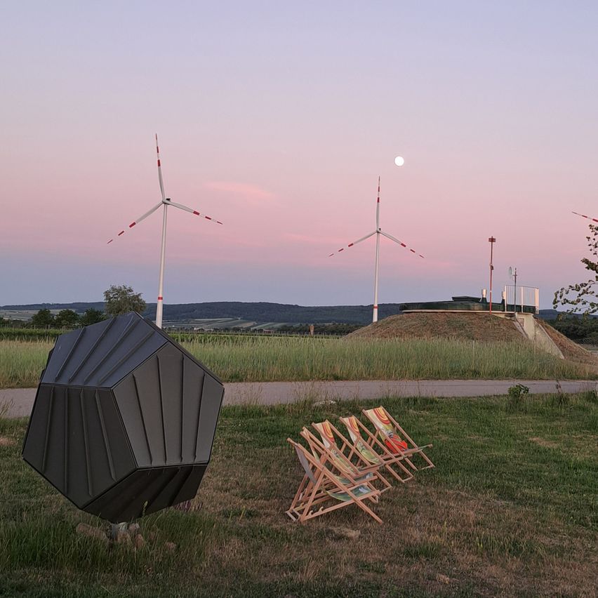 A black geometric object stands in a field with wind turbines in the background. There are four wooden deck chairs nearby. The moon is in the sky.