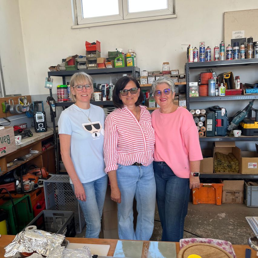 Drei Frauen stehen lächelnd in einer Werkstatt. Hinter ihnen befinden sich Regale mit Werkzeugen, Lagerboxen und einem Fenster. Ein Tisch davor hält ein in Silberfolie eingewickeltes Objekt.