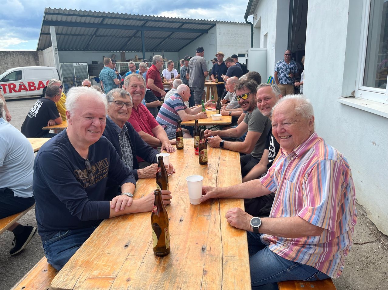 Eine Gruppe von Männern sitzt um einen Holztisch, lächelt, mit Flaschen und Gläsern. Der Tisch hat eine Bierflasche und einen weißen Becher. Sie sind wahrscheinlich in einem Restaurant.