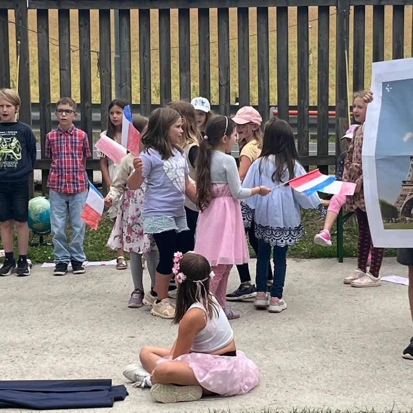 Eine Gruppe von Kindern in Schuluniformen steht und sitzt auf einem Feld und hält Flaggen. Ein Kind sitzt auf dem Boden und hält ein Poster. Ein Globus steht in der Nähe.