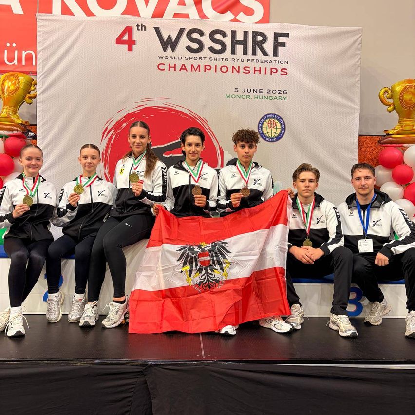 Sechs Athleten stehen bei den 4. WSSHR World Sport Shito Ryu Federation Championships auf der Bühne, halten Medaillen und eine österreichische Flagge und ein Hintergrund zeigt Event-Details.