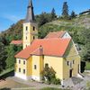 Luftaufnahme einer gelben Kirche mit Turm und rotem Dach, umgeben von viel Grün und einem klaren blauen Himmel.