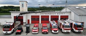 Eine Reihe roter Feuerwehrfahrzeuge, die vor der FF Hafnerbach Mark Feuerwehrstation unter einem bewölkten Himmel geparkt sind, mit zwei Drohnen in der Luft.