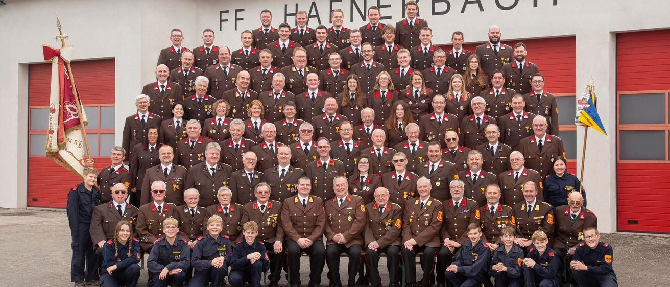 Eine große Gruppe von Feuerwehrleuten und Familienmitgliedern posiert für ein Foto vor der FF Haenerbach Feuerwehrwache, mit verschiedenen Uniformen und Accessoires.
