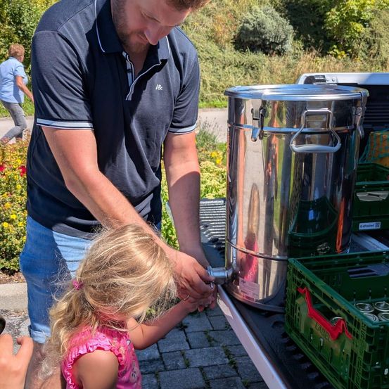 Ein Mann hilft einem kleinen Mädchen, Wasser in einen Metallbehälter zu gießen, in einer Außenumgebung.