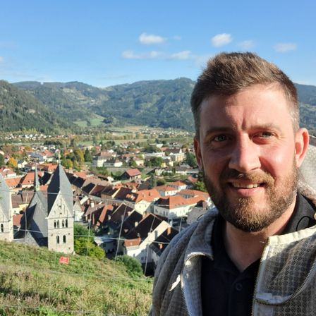 Ein lächelnder Mann steht vor einem Dorf mit orangefarbenen Dächern. Hinter ihm ist ein klarer blauer Himmel und eine Kulisse von Bergen.