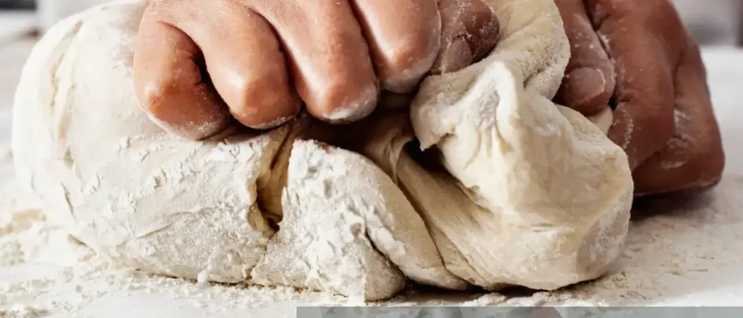 Eine Person in einer weißen Schürze knetet Teig auf einer bemehlten Oberfläche, möglicherweise zur Vorbereitung von Brot.