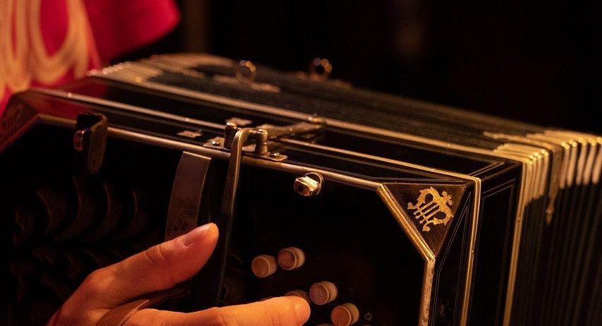Eine Person spielt ein schwarzes Akkordeon mit goldener Einfassung, die Finger drücken auf die Tasten. Ein rotes Hemd ist im Hintergrund zu sehen.