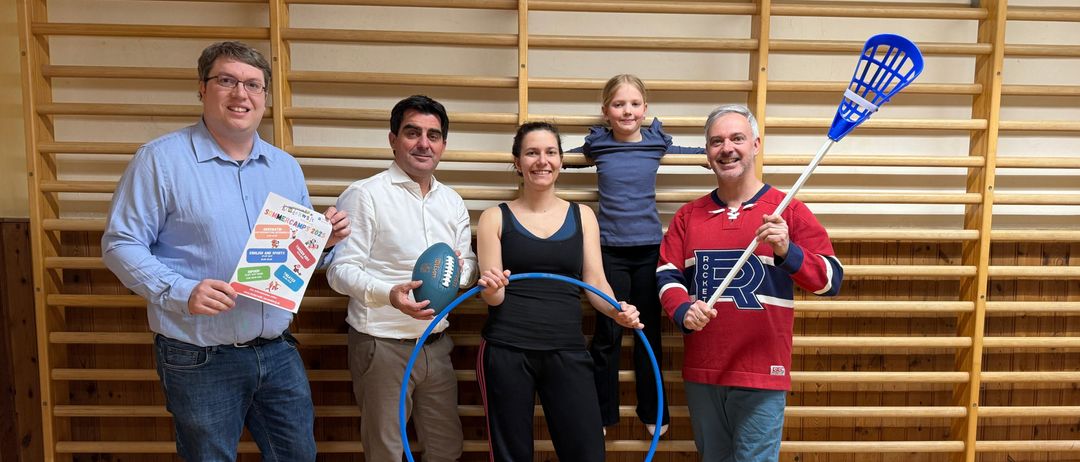 Fünf Personen stehen in einem Raum mit einer hölzernen Wand im Hintergrund. Sie halten verschiedene Sportgeräte, darunter einen Lacrosse-Schläger, einen Fußeball und eine Hula-Hoop.