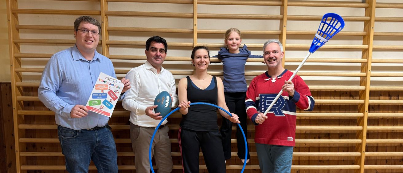 Fünf Personen stehen in einem Raum mit einer hölzernen Wand im Hintergrund. Sie halten verschiedene Sportgeräte, darunter einen Lacrosse-Schläger, einen Fußeball und eine Hula-Hoop.