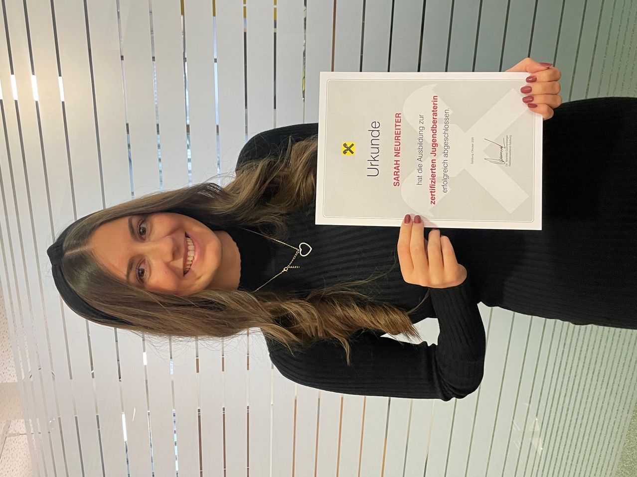 Eine lächelnde Frau hält ein Zertifikat für Sarah Neureiter, das ihr Alter für Jugendangelegenheiten bestätigt. Sie trägt ein schwarzes Oberteil und hält das Zertifikat vor einem weiß gerahmten Fenster.