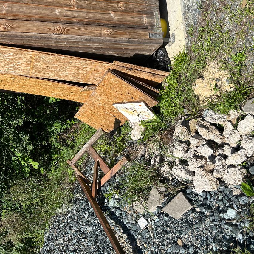 Eine Draufsicht zeigt einen Haufen zerbrochener Holzbretter und eine Tafel mit Schriftzügen, verstreut auf Kies und Gras.