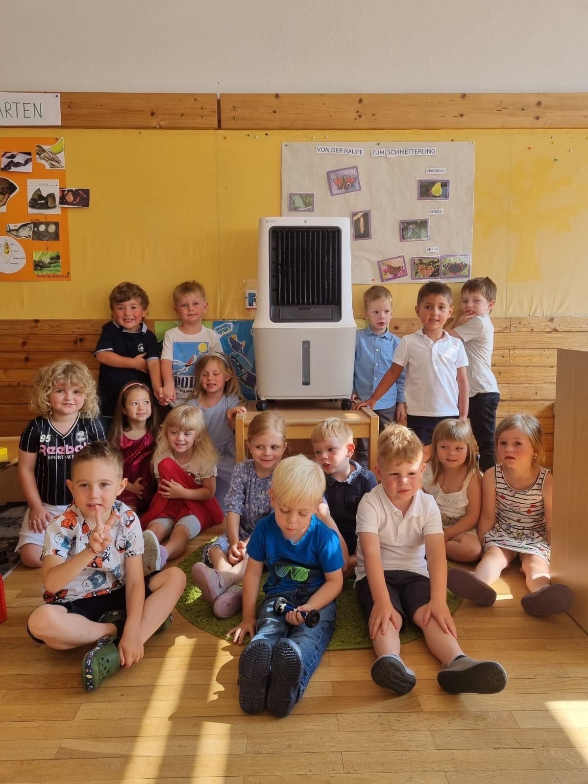 Ein Klassenzimmer mit einer Gruppe von Kindern, die auf dem Boden um einen weißen Luftkühler sitzen, eine gelbe Wand mit Lehrplakaten und eine Holzwand im Hintergrund.