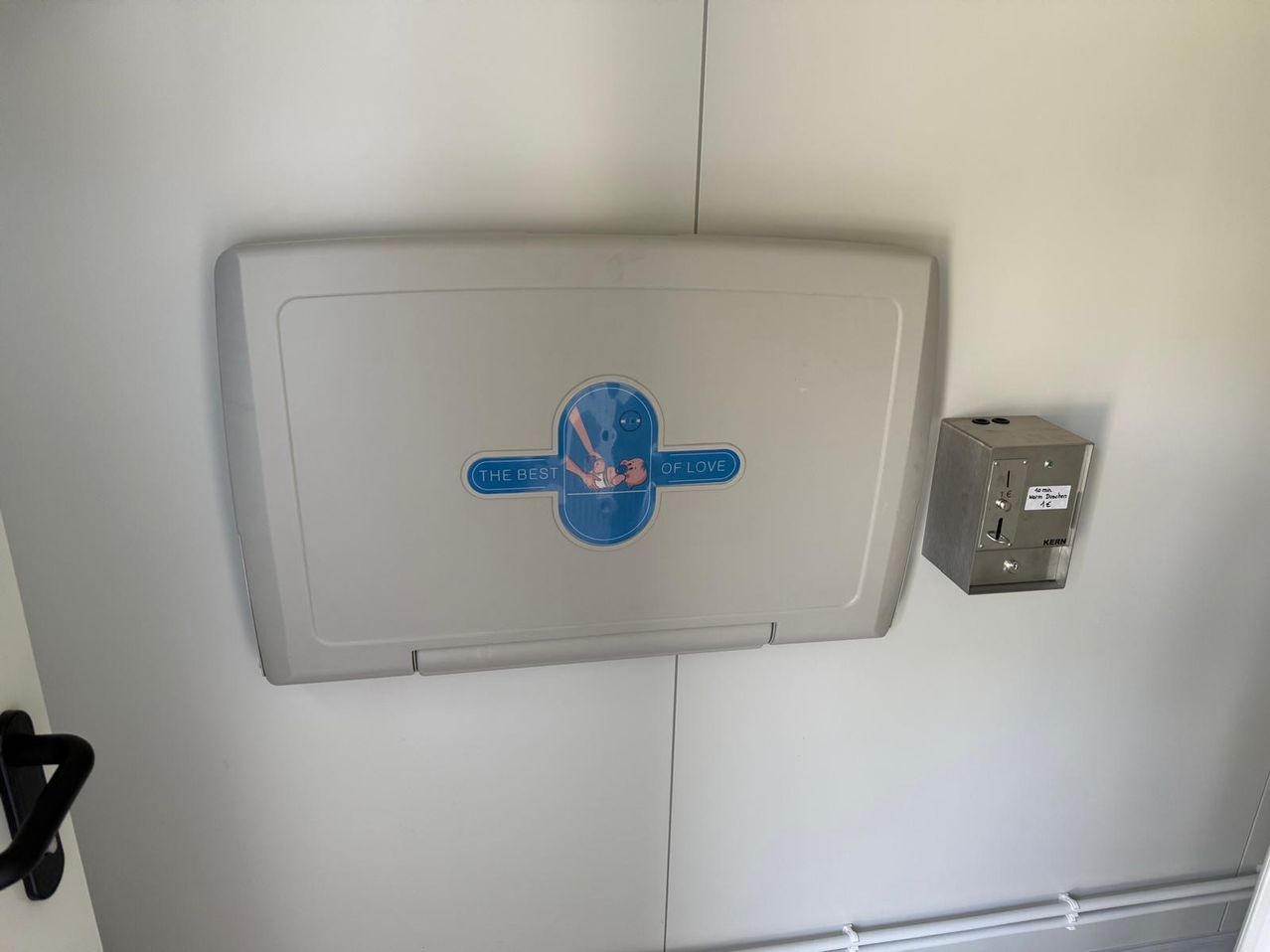 Eine Wickelstation mit einem blauen Logo, das 'The Best of Love' steht, ist an einer weißen Wand montiert. Daneben befindet sich eine Metallbox mit einem kleinen Aufkleber.