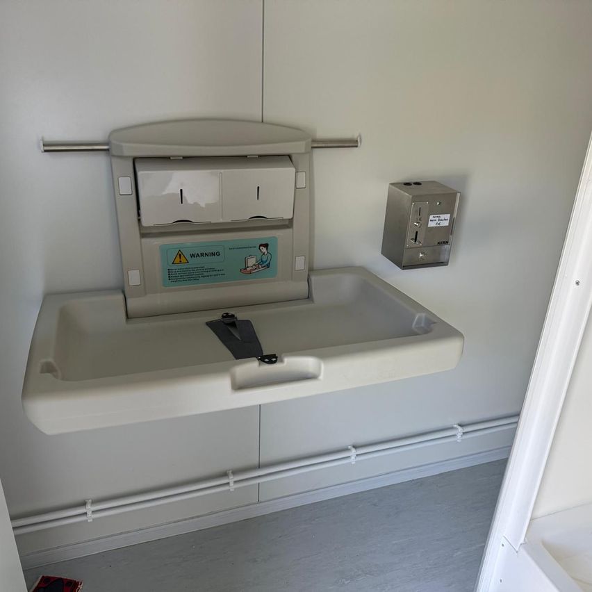 Eine Baby-Windelstation ist an einer weißen Wand montiert, einschließlich eines Beckens und eines Warnhinweises. In der Nähe befindet sich ein Spender.