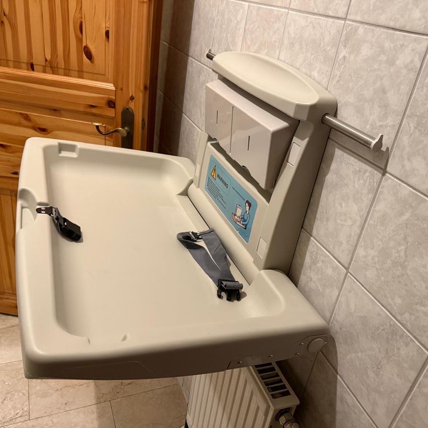 Ein weißes Wickeltisch mit Ablage, Sicherheitsgurt und Warnaufkleber ist an einer gefliesten Wand montiert. Dahinter befindet sich eine Holztür und ein Heizkörper.