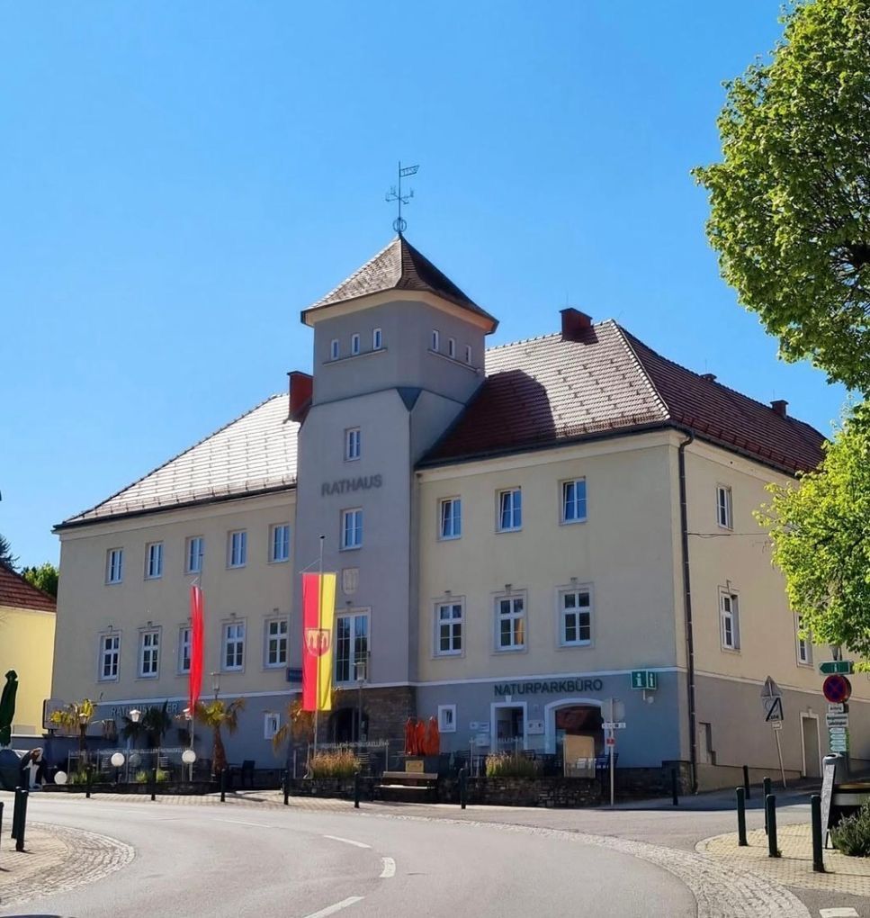 Das Rathaus, ein großes Gebäude mit einem braunen Dach, verfügt über einen Turm mit einer Wetterfahne und Flaggen. Das Gebäude hat viele Fenster und ist beige gestrichen. Vor dem Gebäude steht ein Schild mit der Aufschrift Naturparkburo.