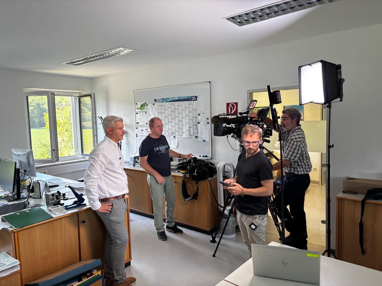 Vier Männer sind in einem Innenstudio. Drei von ihnen stehen und einer bedient eine Kamera. Sie sind von Ausrüstung und einer Tafel umgeben.
