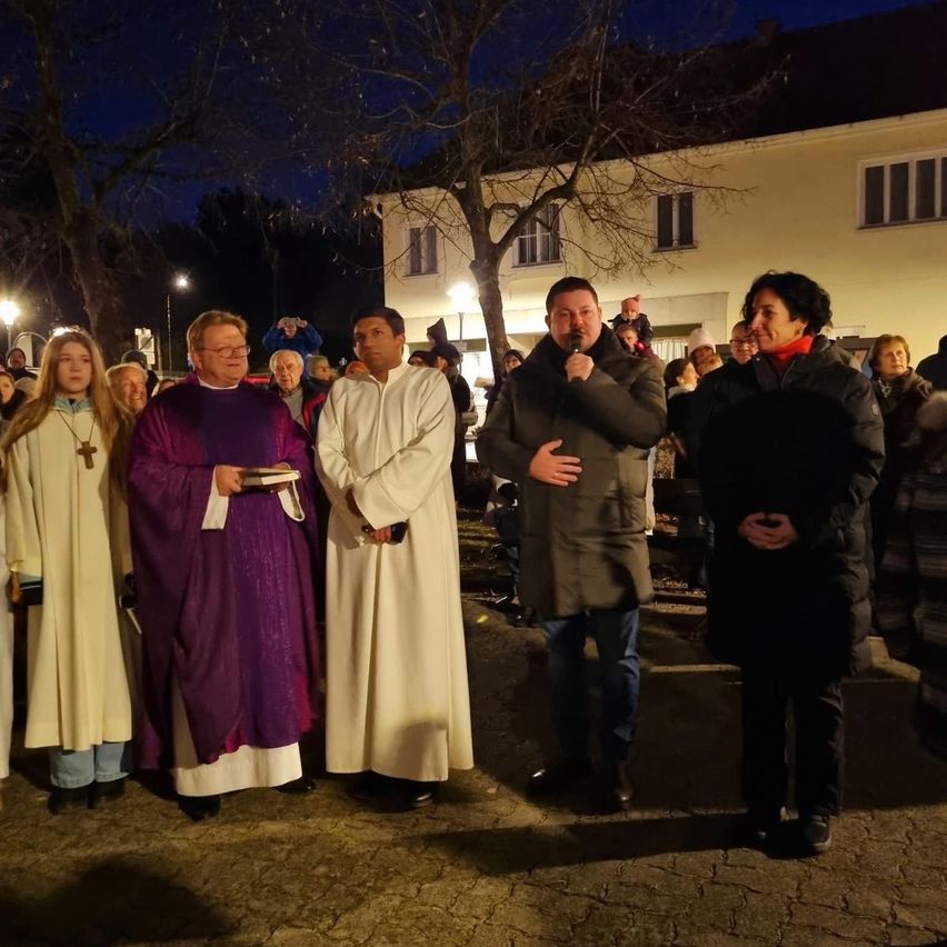 In der Nacht stehen eine Gruppe von Menschen, einschließlich Priester, zusammen vor einem Haus. Ein Mann hält ein Mikrofon und scheint zu sprechen.