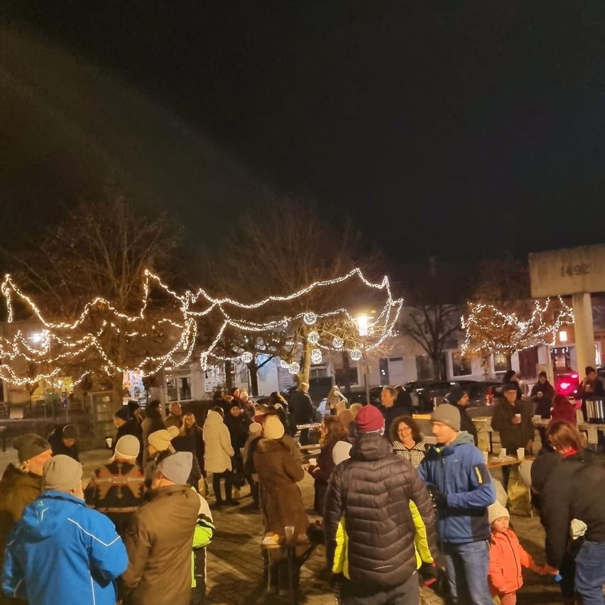 Eine Menschenmenge versammelt sich im Freien unter einem Nachthimmel, geschmückt mit Lichterketten. Einige tragen Winterkleidung und Mützen, während andere in der Nähe eines Tisches mit Getränken stehen. Bäume und Gebäude im Hintergrund sind von den Lichtern beleuchtet.