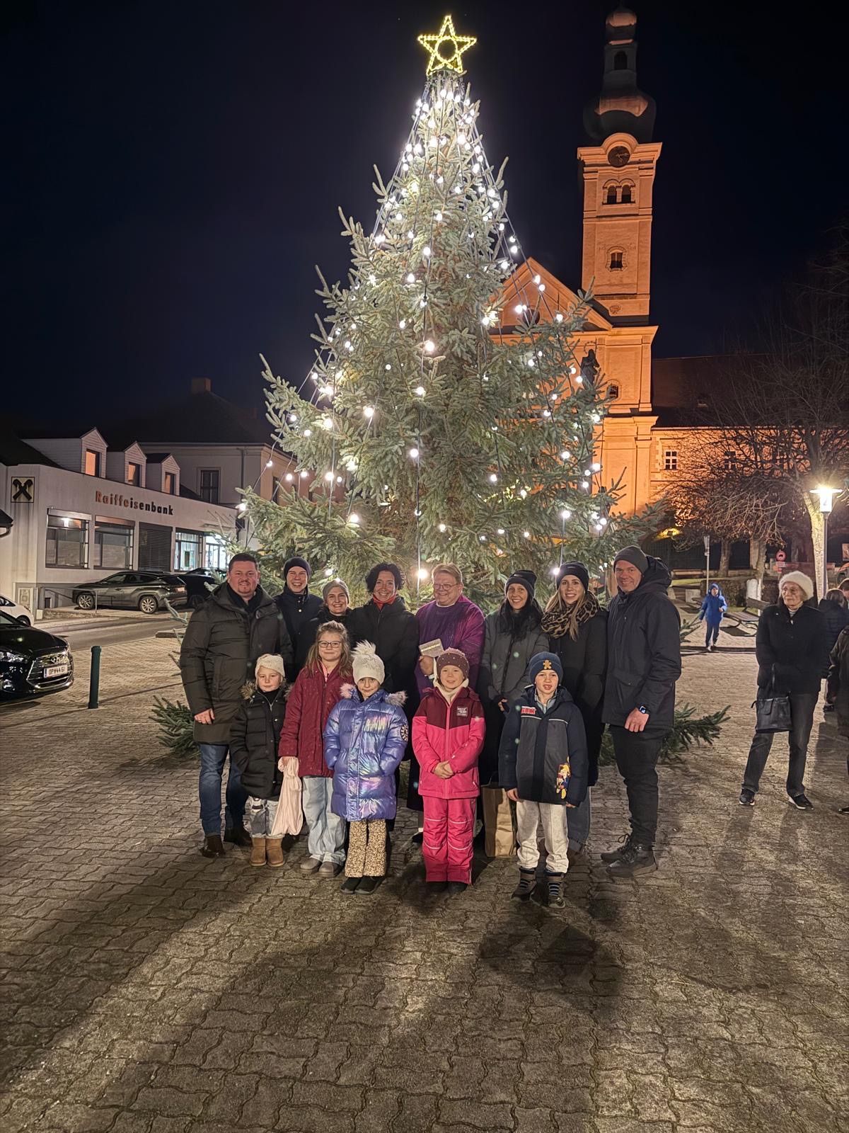 Eine Gruppe von Menschen steht vor einem großen Weihnachtsbaum und posiert für ein Foto bei Nacht. Dahinter ist eine Kirche mit einem Turm und einem beleuchteten Schild zu sehen.