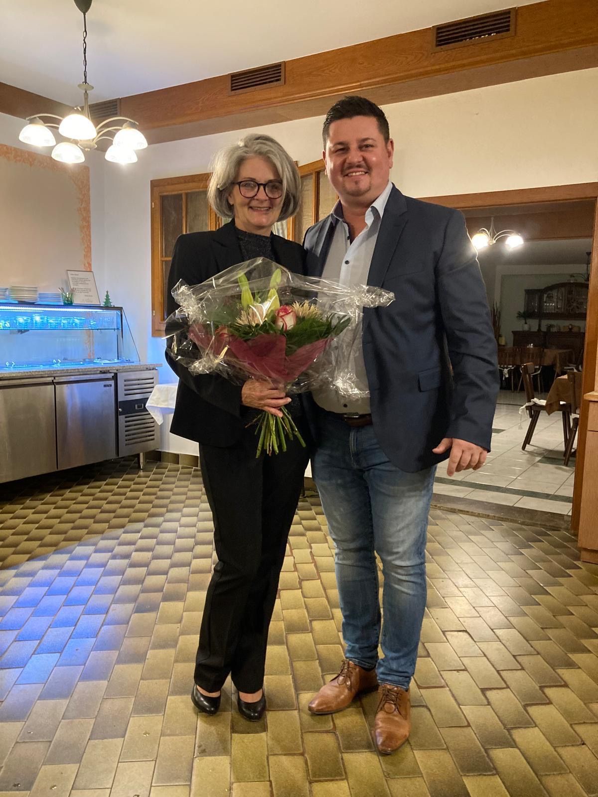 Eine Frau in einem schwarzen Anzug hält einen Blumenstrauß und steht neben einem Mann in einem dunkelblauen Anzug. Sie befinden sich in einem Raum mit gefliestem Boden, einer Küchenecke im Hintergrund und einer lächelnden Frau.