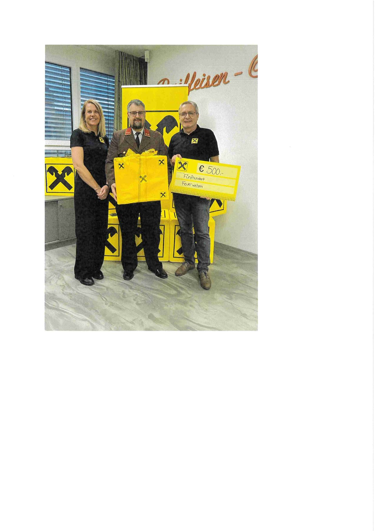 Drei Personen stehen in einem Raum, zwei Männer und eine Frau, halten gelbe Objekte. Der Mann auf der rechten Seite hält einen Scheck, und der in der Mitte hält eine Geschenkbox.