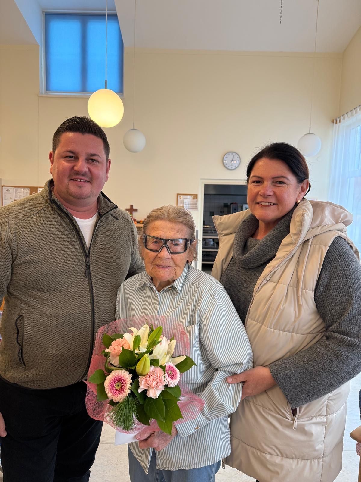 Ein Mann und eine Frau lächeln für ein Foto, neben einer älteren Frau, die einen Blumenstrauß hält, in einem Raum mit einer weißen Wand und hängenden Lampen.