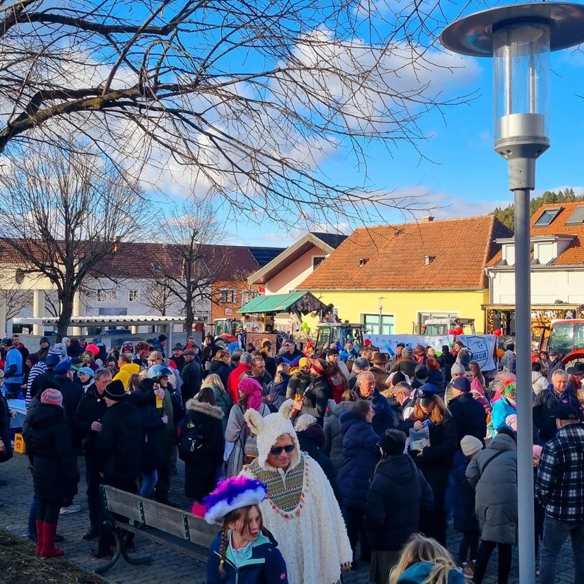 A large crowd gathers outdoors, with some individuals wearing winter attire and others in costumes. A girl in a pink hat stands beside a person in a white furry costume. Buildings and a tree are visible in the background.