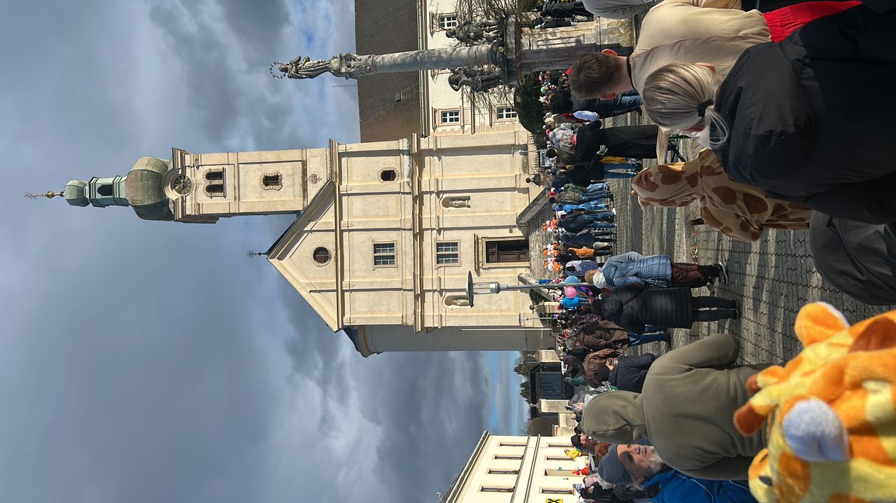 A crowd gathers in front of a large, ornate church building with tall towers, under a cloudy sky.