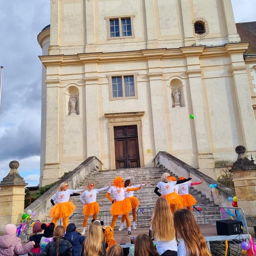 A group of women dressed in orange tutus are performing on a stage in front of a church, with spectators watching.