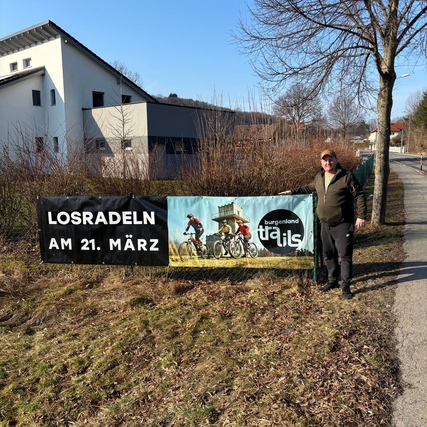 A man stands next to a banner advertising a biking event called Losradeln in Burgenland, with a building and trees in the background.