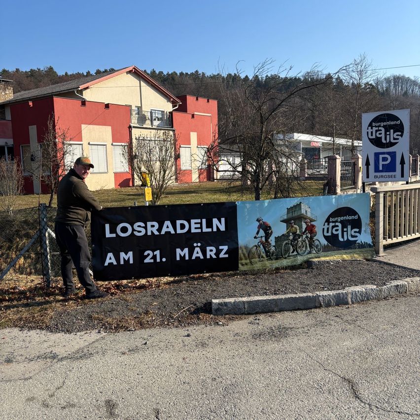 A man stands by a banner advertising an event named Losradeln on 21st March. There are bicycles on the banner, with trees and a building in the background.