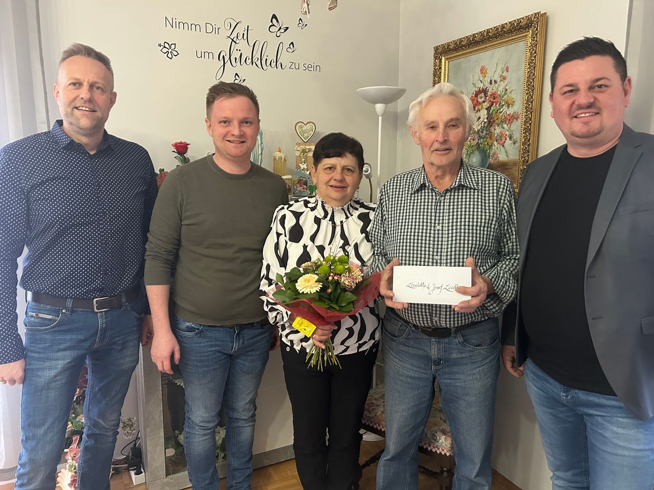 Five people stand together in a room. A woman holds a bouquet of flowers. A man holds a card. Behind them is a wall with text.