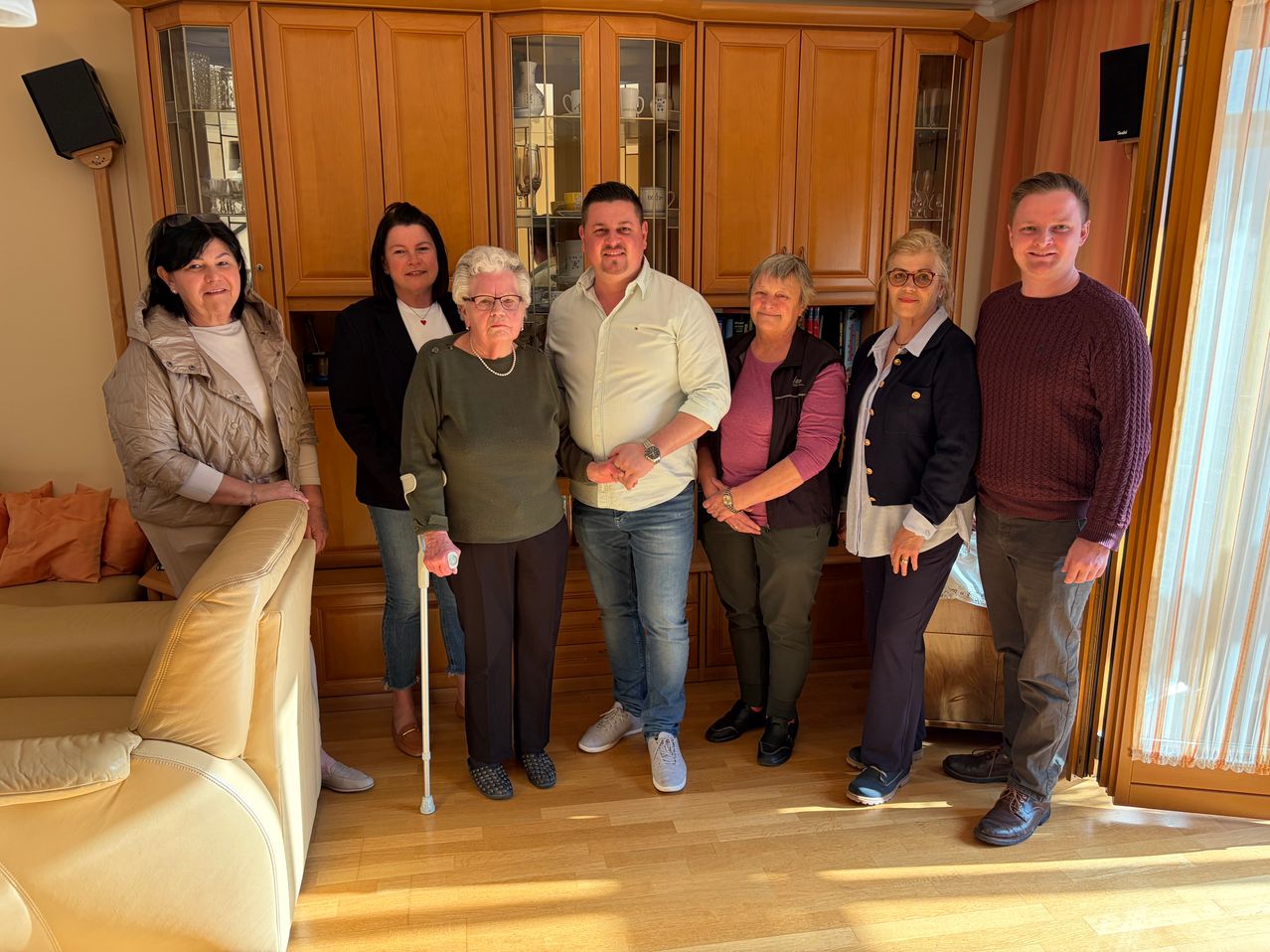 Eine Gruppe von sieben Personen posiert für ein Foto im Wohnzimmer, auf einem Holzboden stehend, mit einem Sofa und Schränken im Hintergrund.