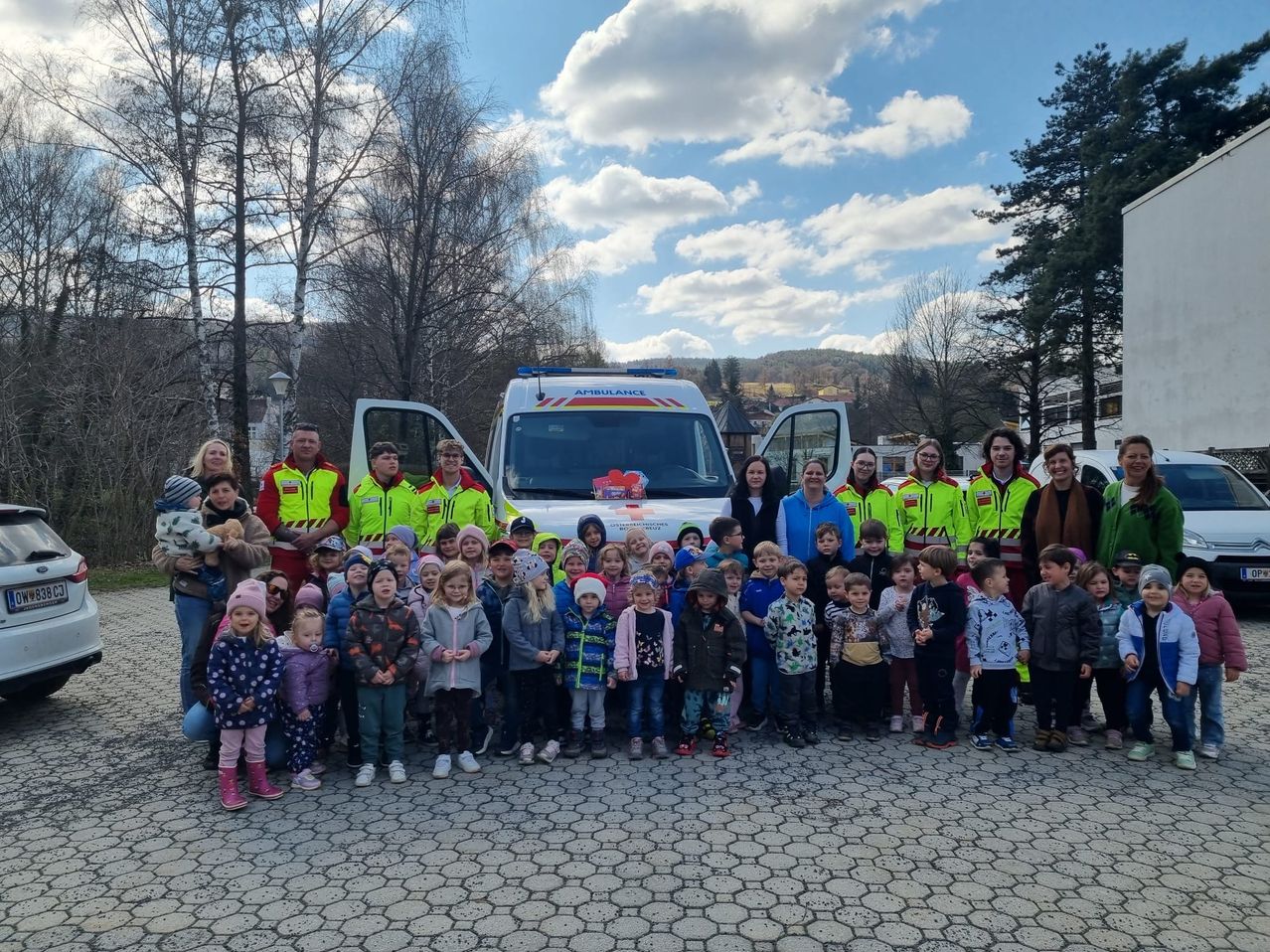 Eine Gruppe von Kindern, Erwachsenen und Rettungsdienstpersonal posiert für ein Foto vor einem Krankenwagen. Bäume und ein bewölkter Himmel sind im Hintergrund zu sehen.