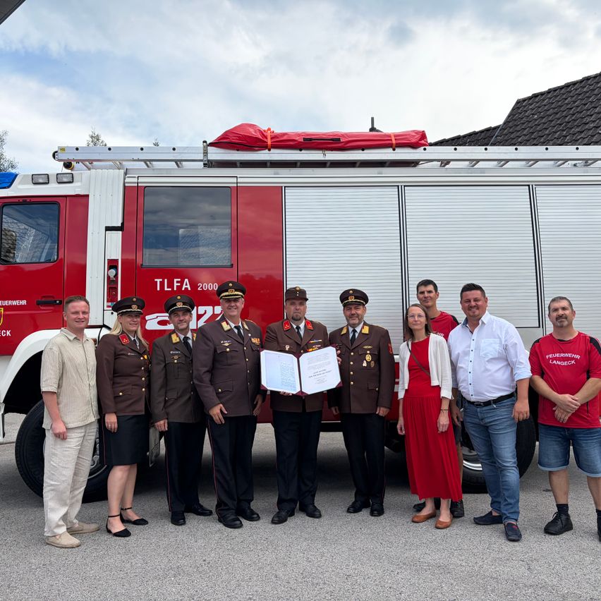 A group of people, including firefighters and civilians, stand in front of a fire truck. They are holding a certificate and appear to be celebrating an achievement.