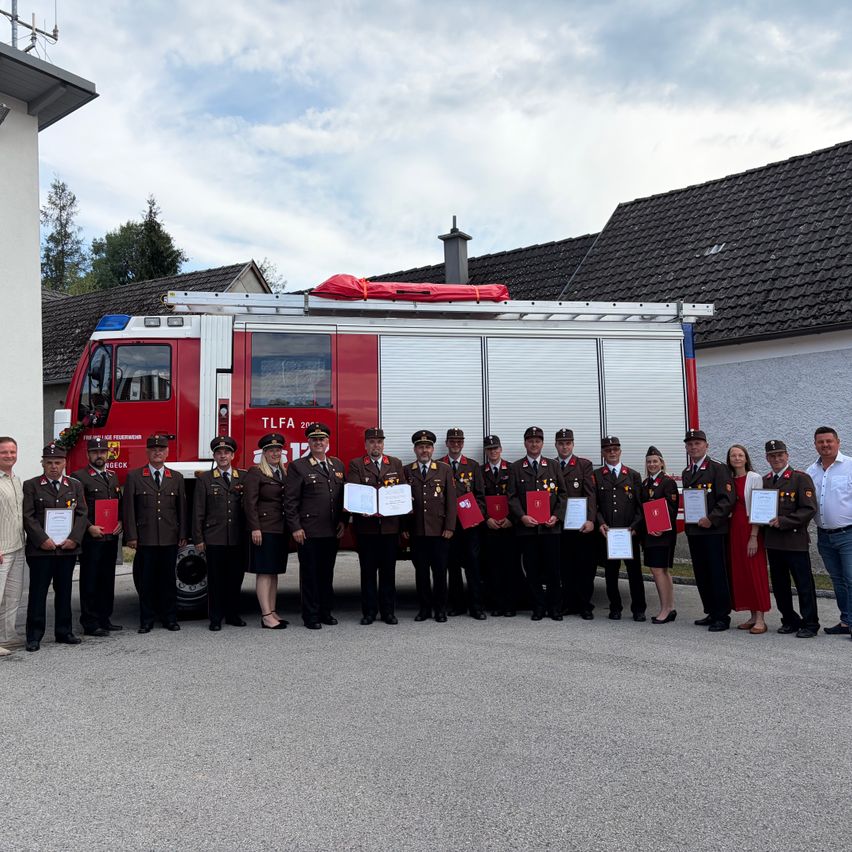 A group of firefighters in uniform stands in front of a fire truck with a red cover on top. They are holding certificates and posing for a photo.