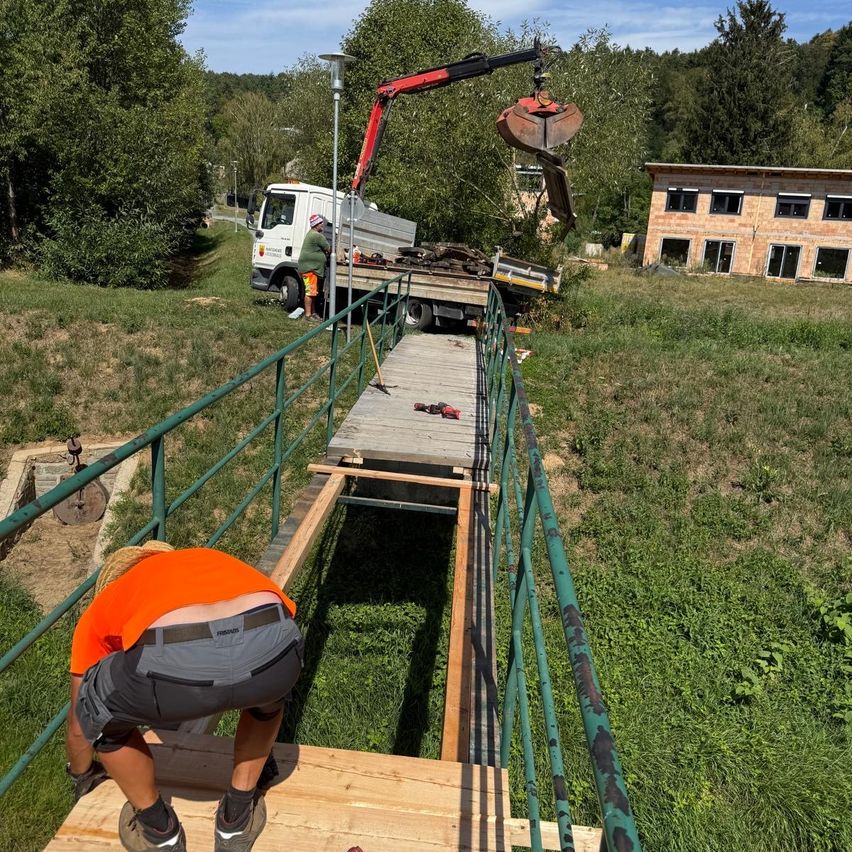 Eine Baustelle mit Arbeitern, einer Person, die sich über eine Holzplattform beugt, einem Kran, der eine Last hebt, und einem Gebäude im Hintergrund.