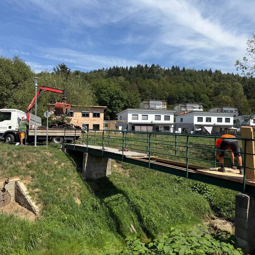 Eine Baustelle mit Arbeitern, die eine Brücke über ein Grasfeld installieren, einem Lastwagen mit einem Kran in der Nähe und mehreren Häusern im Hintergrund.