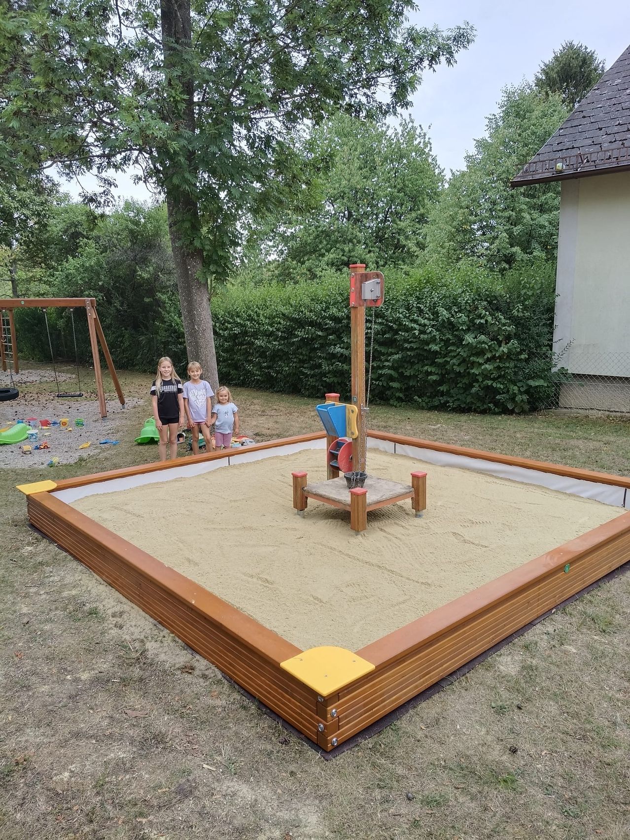 Three young girls stand near a wooden sandbox with a play structure in a backyard.
