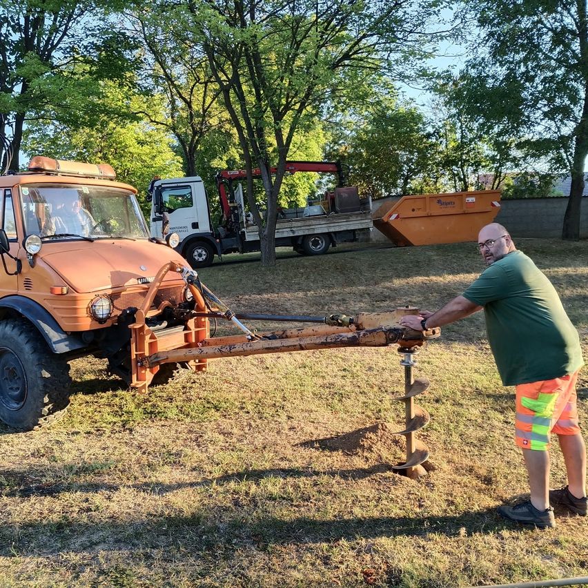 A man in a green shirt and orange shorts is operating a large orange drilling machine on a grassy field. Behind him, a white truck is parked, and a large orange dumpster is nearby. Trees and a building are in the background.
