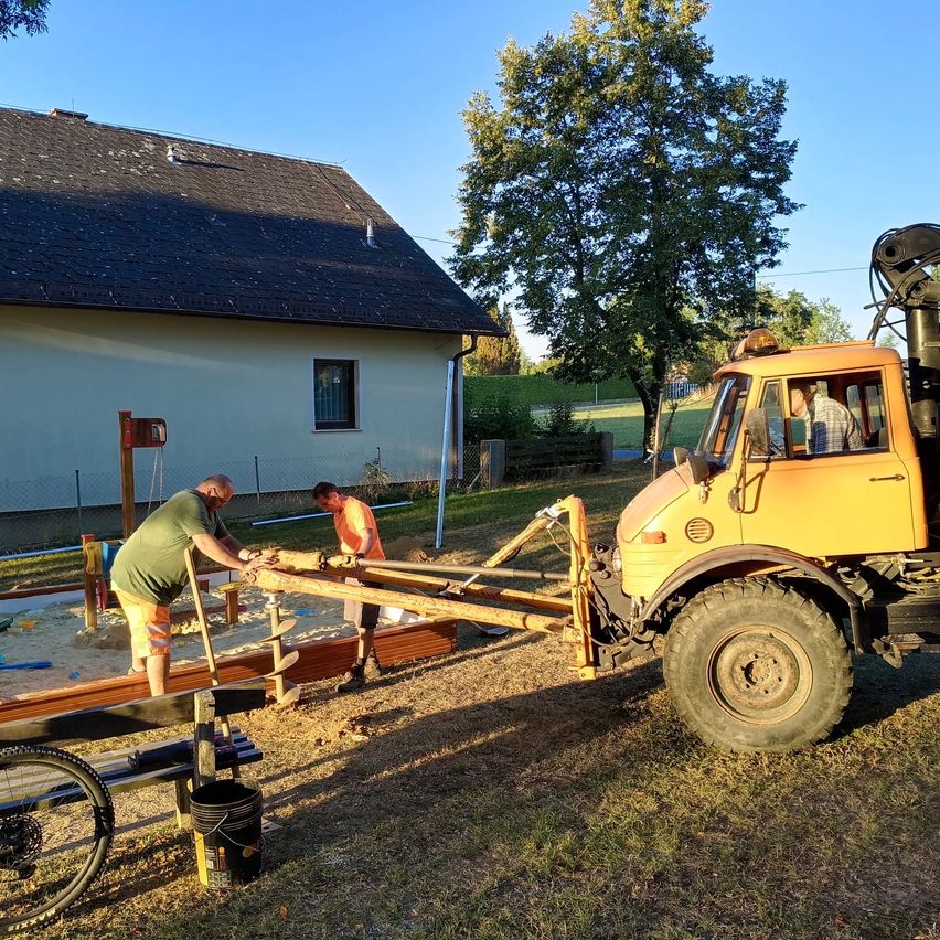 Two men are working on a wooden structure in a yard, with a yellow machinery vehicle nearby. A house with a black roof is in the background, and a tree is in front of it.