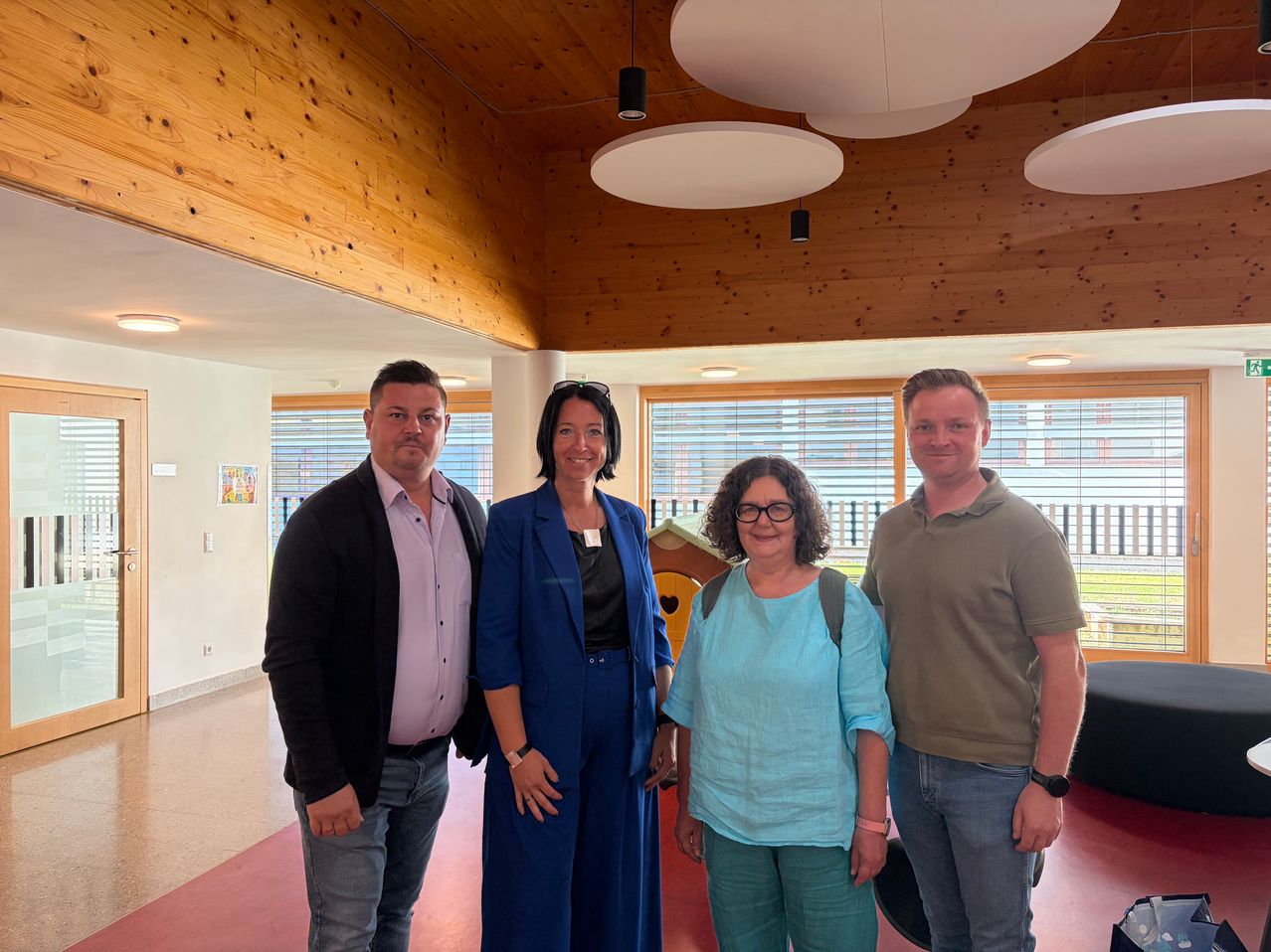 Four individuals, two men and two women, stand together in a room with wooden walls and large windows. The woman in the middle wears a blue blazer, and the man on the left wears a black jacket.
