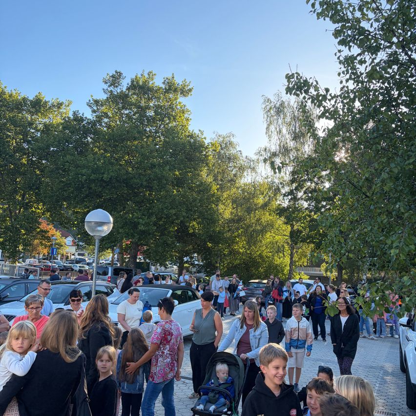 A crowd of people, including families with children, are gathered in a park with trees and parked cars. Some are walking, while others are standing. A stroller with a baby is visible in the foreground.