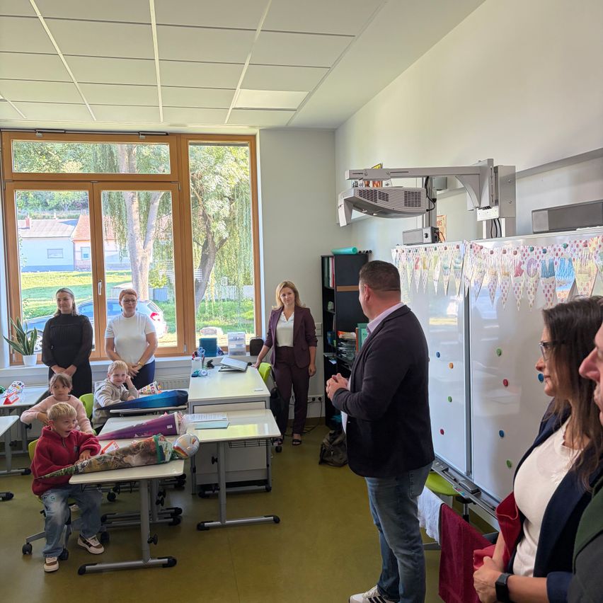 A group of people in a classroom, with a man speaking in front of a whiteboard. Children sit at desks, and adults stand around. Outside, a tree and building are visible through the windows.