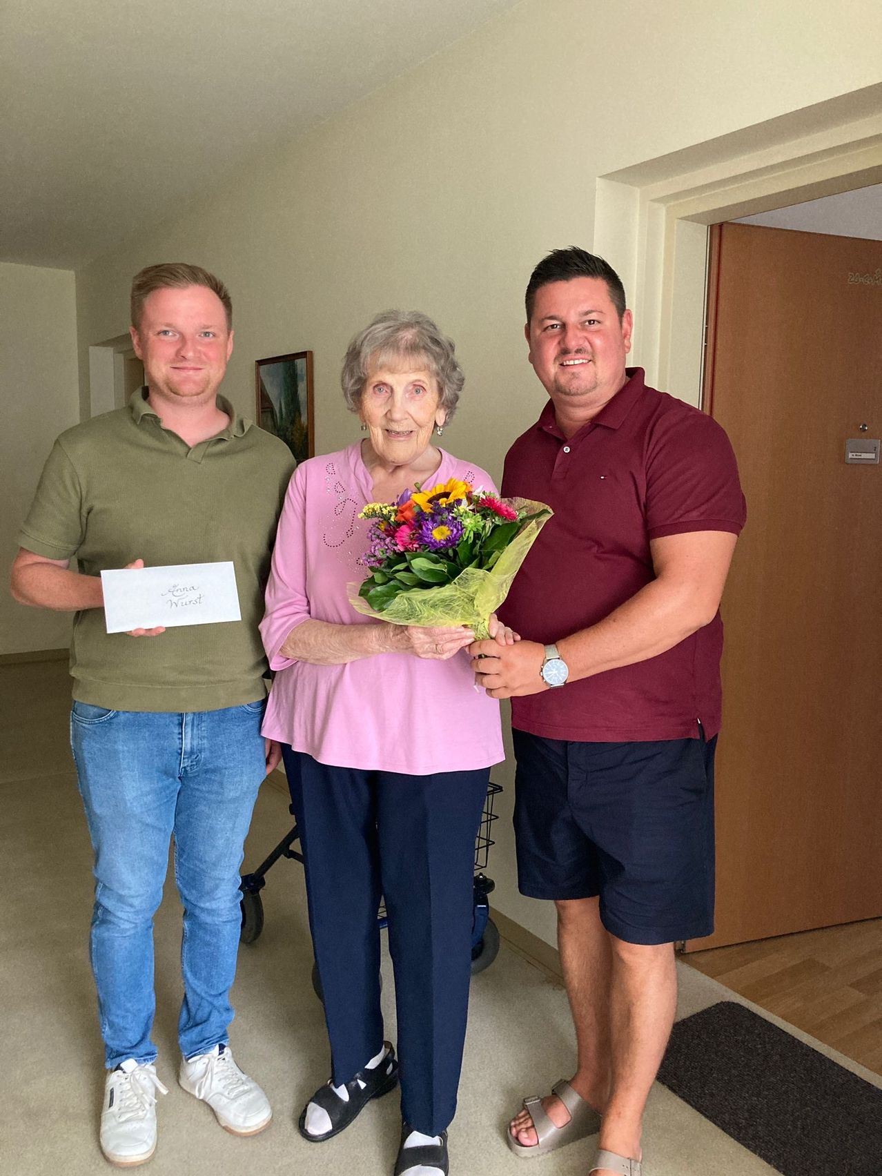 An elderly woman holding a bouquet of flowers stands between two smiling men in a room.
